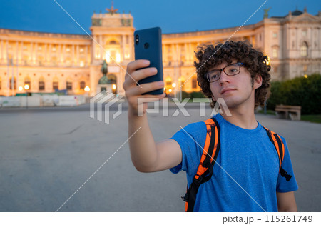 Vienna, Austria, August 17, 2022. A young Caucasian man with curly hair and glasses takes a selfie in front of the Hofburg Imperial Palace at sunset. The warm evening light enhances the imposing 115261749