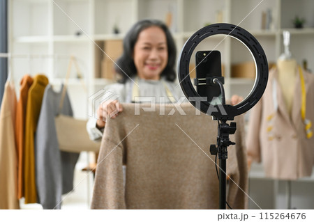 Smiling middle age woman showing clothes in front of smartphone camera while live streaming at her shop 115264676