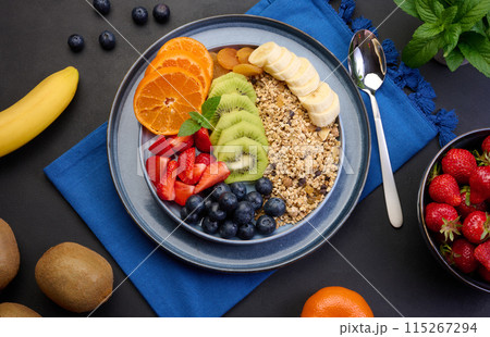 Granola with strawberries, kiwi, banana and blueberries in a round plate on a black table. Healthy and tasty food Granola with strawberries, kiwi, banana and blueberries in a round plate on a black table. Healthy and tasty food 115267294