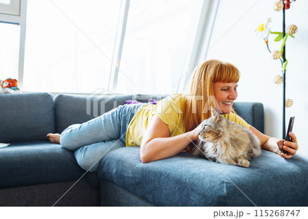 portrait positive blonde middle-aged woman lying on sofa hugging cat taking selfie portrait positive blonde middle-aged woman lying on sofa hugging cat taking selfie 115268747