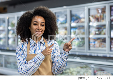 A cheerful supermarket employee in an apron, smiling and pointing at products in the frozen food aisle. Retail and customer service concept. A cheerful supermarket employee in an apron, smiling and pointing at products in the frozen food aisle. Retail and customer service concept. 115270951