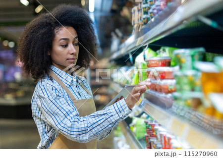 Young female grocery store worker using a tablet to manage inventory in an aisle filled with various products. Young female grocery store worker using a tablet to manage inventory in an aisle filled with various products. 115270960