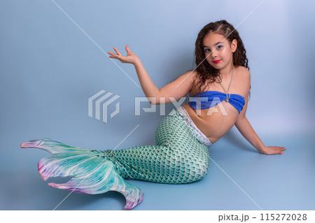 A beautiful elegant girl in a mermaid costume lies on a blue background with her palm raised up. 115272028