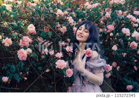 Fairy tale woman portrait. Young beautiful woman Fairy holds pink flower rose on blooming bush background. Hairstyle loose green hair, romantic purple medieval vintage dress. Summer fantasy garden. Fairy tale woman portrait. Young beautiful woman Fairy holds pink flower rose on blooming bush background. Hairstyle loose green hair, romantic purple medieval vintage dress. Summer fantasy garden. 115272345