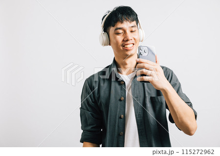 An excited Asian man, with a happy smile, enjoys karaoke on his smartphone using Bluetooth headphones for a wireless experience. Studio shot isolated on white background, capturing his positive vibes. 115272962