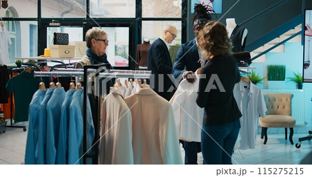 Fashion stylist suggesting clothing items to regular customer, team of store workers help elderly woman to buy trendy clothes on sale. Retail clerks assisting client during shopping spree. Camera B. Fashion stylist suggesting clothing items to regular customer, team of store workers help elderly woman to buy trendy clothes on sale. Retail clerks assisting client during shopping spree. Camera B. 115275215