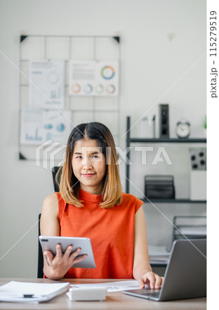 A woman in an orange shirt is sitting at a desk with a tablet and laptop in front of her. She is holding a tablet in her hand and she is working on a project. The room is filled with papers and books A woman in an orange shirt is sitting at a desk with a tablet and laptop in front of her. She is holding a tablet in her hand and she is working on a project. The room is filled with papers and books 115279519