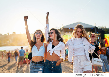 Young women dance at sunset on sandy shore, enjoying music festival. Friends celebrate, summer beach party vibe, arms uplifted in joy. Casual fashion, sea backdrop, golden hour light at music event. 115284822