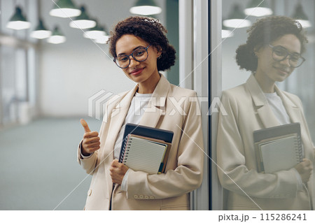 Pretty female manager in eyeglasses standing on office background and showing thumb up 115286241