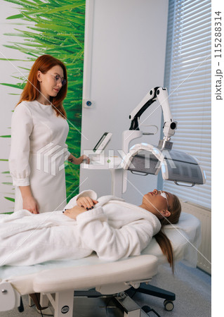 Vertical shot of professional beautician specialist connecting and adjusting device for photo peeling at modern aesthetic clinic. Woman lying during non-invasive type of phototherapy. Vertical shot of professional beautician specialist connecting and adjusting device for photo peeling at modern aesthetic clinic. Woman lying during non-invasive type of phototherapy. 115288314