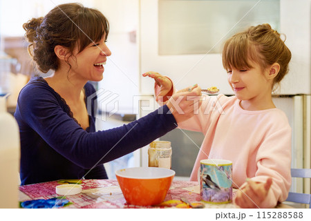 Happy mother, child and feeding with cereal for fiber, vitamin or breakfast in kitchen together at home. Mom, little girl or young daughter with spoon of porridge for healthy meal, nutrition or snack 115288588