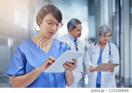 Woman, doctor and tablet in hospital with scrubs for medical, check on digital file for schedule. Female surgeon, medicine and tech with colleagues in background and working, career in healthcare 115288688
