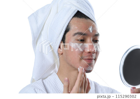 Young man applying moisturizer on his face and looking at mirror over white background Young man applying moisturizer on his face and looking at mirror over white background 115290302