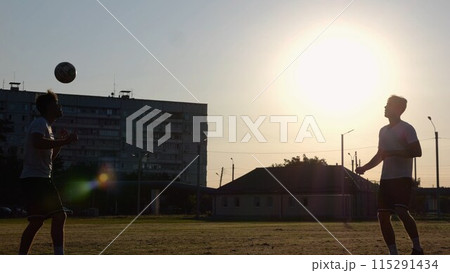 Silhouette of professional footballers kicking soccer ball with their heads on stadium at sunset. Two sportsmen showing tricks with ball while passing it to each other at field. Freestyle football 115291434