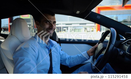 Profile of handsome businessman driving a modern electric car at urban road. Male successful entrepreneur riding on electrical vehicle at city. Young man commuting to work at sunny day. Slow motion 115291458