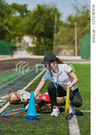 Female coach preparing for outdoor soccer training for children. Arranging necessary equipment for training and warming up. Female coach preparing for outdoor soccer training for children. Arranging necessary equipment for training and warming up. 115296456