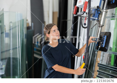 Female operator maneuvering glass sheets with vacuum lifter in workshop 115301676