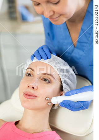 Adult woman cosmetologist doing microcurrent procedure to young woman Adult woman cosmetologist doing microcurrent procedure to young woman 115302143