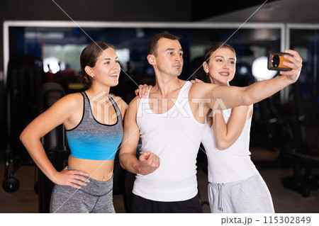 Two cheerful girls and young man taking selfie in fitness hall 115302849