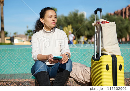 Woman sitting outdoors with luggage and cell phone, waiting for transport or enjoying a break Woman sitting outdoors with luggage and cell phone, waiting for transport or enjoying a break 115302901