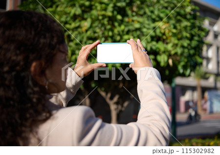 View from the back to a woman holding smartphone with white blank mockup screen, taking photo of a city on sunny day 115302982