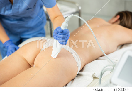 Adult female cosmetologist performs scrubbing procedure on butt of young woman 115303329