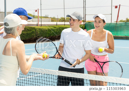 Group of tennis players standing on open court on warm day with rackets and balls in hands, friendly chatting after match 115303963