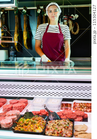 Friendly female butcher behind meat preparations display counter 115304127