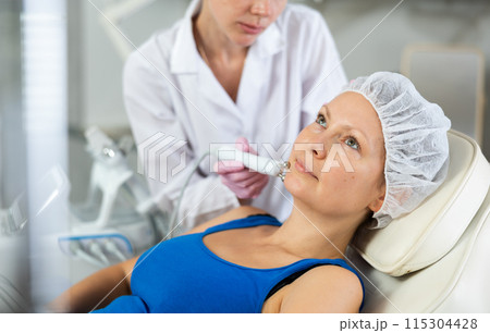 Woman getting facial radio wave lifting skin tightening procedure in salon. RF-lifting device treatment of acne scars, cellulite, skin rejuvenation, Concept of professional cosmetology and Woman getting facial radio wave lifting skin tightening procedure in salon. RF-lifting device treatment of acne scars, cellulite, skin rejuvenation, Concept of professional cosmetology and 115304428