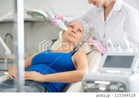 Young female cosmetologist doing exfoliating procedure to adult woman Young female cosmetologist doing exfoliating procedure to adult woman 115304529