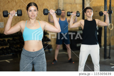 Young dedicated active strong fit woman doing exercises for shoulders while pushing up dumbbells in CrossFit center Young dedicated active strong fit woman doing exercises for shoulders while pushing up dumbbells in CrossFit center 115305639
