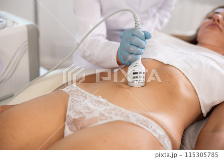 Cosmetologist performing cryotherapy using cooling attachment on female client belly 115305785
