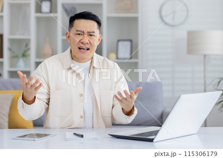 Man expressing frustration during a video call meeting at home. He is seated at a desk with a laptop and paperwork. Man expressing frustration during a video call meeting at home. He is seated at a desk with a laptop and paperwork. 115306179