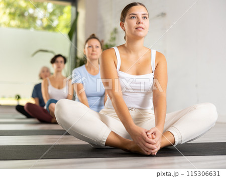 Concentrated young woman performing Butterfly Pose Baddha Konasana during yoga class with female group Concentrated young woman performing Butterfly Pose Baddha Konasana during yoga class with female group 115306631