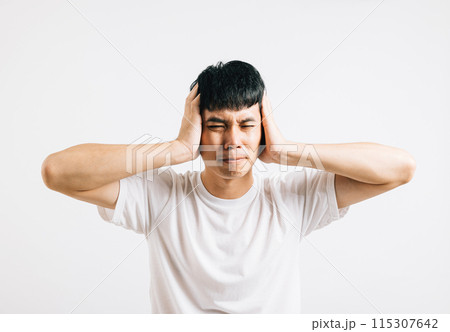 An Asian man in pain, using his hands to cover his ears and shutting his eyes to cope with the loud noise. Studio shot isolated on white background, showing his need for relief. 115307642