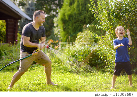 Funny little boy with his father playing with garden hose in sunny backyard. Preschooler child having fun with spray of water. Summer vacation in the village for kids. 115308697