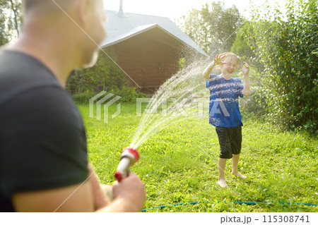 Funny little boy with his father playing with garden hose in sunny backyard. Preschooler child having fun with spray of water. Summer outdoors activity for kids. 115308741