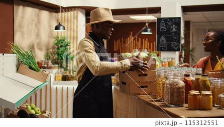 Local vendor placing crates with freshly harvested fruits on grocery store display, recommending organic produce to regular customer. African american farmer restocking all merchandise. Camera 2. 115312551