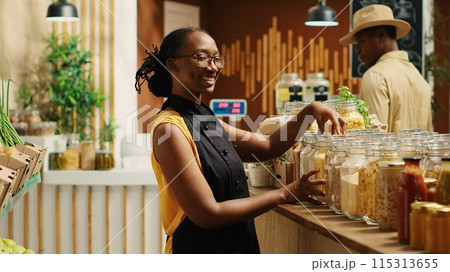 Local merchant organizing homemade pasta in reusable jars at zero waste supermarket, wearing apron and welcoming customers. African american woman in eco friendly store. Camera 1. Local merchant organizing homemade pasta in reusable jars at zero waste supermarket, wearing apron and welcoming customers. African american woman in eco friendly store. Camera 1. 115313655