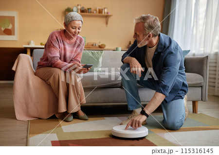 Portrait of senior man pressing on button on robot vacuum cleaner with modern mature woman setting up smart home devices via smartphone copy space Portrait of senior man pressing on button on robot vacuum cleaner with modern mature woman setting up smart home devices via smartphone copy space 115316145