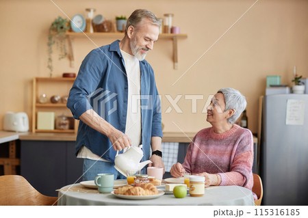 Front view portrait of happy senior couple enjoying breakfast together in cozy kitchen with smiling husband pouring tea copy space 115316185