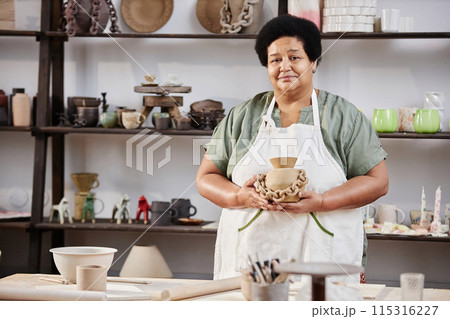 Waist up portrait of senior Black woman smiling at camera and holding handmade pottery piece in workshop, copy space 115316227