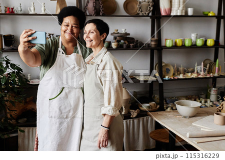 Portrait of two mature women taking selfie photo together in pottery studio, copy space 115316229