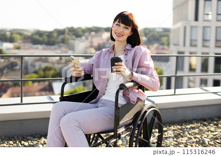 Smiling woman in wheelchair holding coffee and smartphone outdoors 115316246