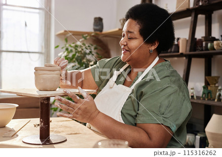 Side view portrait of senior African American woman enjoying pottery class in cozy art studio and hand shaping ceramic mug copy space 115316262