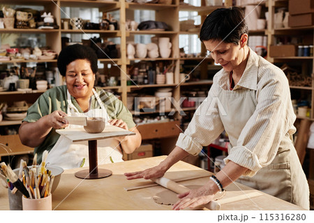 Sunlit portrait of two creative senior women enjoying pottery class in retirement and shaping clay copy space 115316280