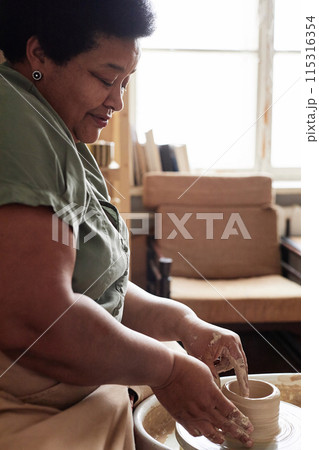 Vertical side view portrait of Black senior woman enjoying pottery class and smiling Vertical side view portrait of Black senior woman enjoying pottery class and smiling 115316354