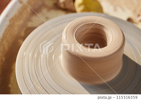 Minimal close up of carefully shaped raw clay on pottery wheel lit by sunlight copy space 115316359
