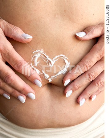 Close up of womans hands middle age with white nails applying cream in the shape of heart on her stomach. Skincare, self care, and personal hygiene with on love and body positivity. Close up of womans hands middle age with white nails applying cream in the shape of heart on her stomach. Skincare, self care, and personal hygiene with on love and body positivity. 115316688
