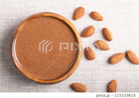 Brown almond butter in a wooden bowl on linen fabric. Smooth food paste made from grinding almonds into nut butter. Puree of nuts of Prunus dulcis with a nutty flavor. A good source of protein. 115319618